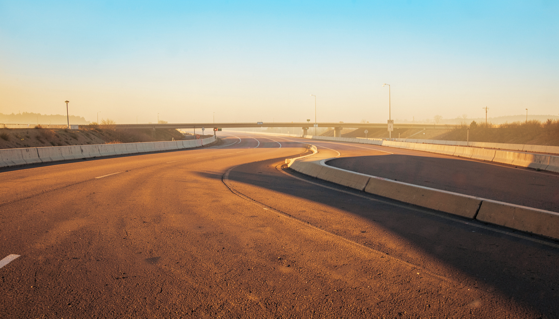 Warm golden-hour photograph of an empty road interchange, glowing amber asphalt and pale concrete curves fading into a soft, hazy horizon with a calm, atmospheric feel.
